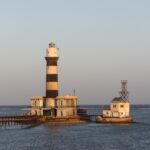 O farol de Daedalus, uma torre listrada de preto e branco sobre o recife, com seus prédios e o píer metálico, ao pôr do sol