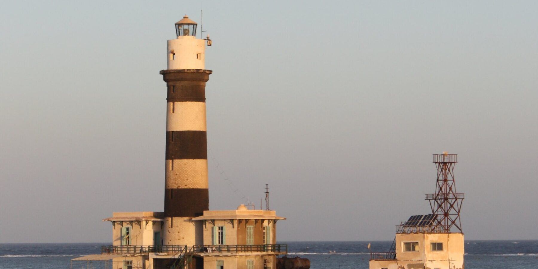 O farol de Daedalus, uma torre listrada de preto e branco sobre o recife, com seus prédios e o píer metálico, ao pôr do sol