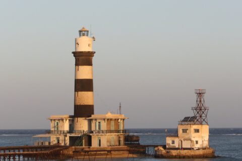 O farol de Daedalus, uma torre listrada de preto e branco sobre o recife, com seus prédios e o píer metálico, ao pôr do sol