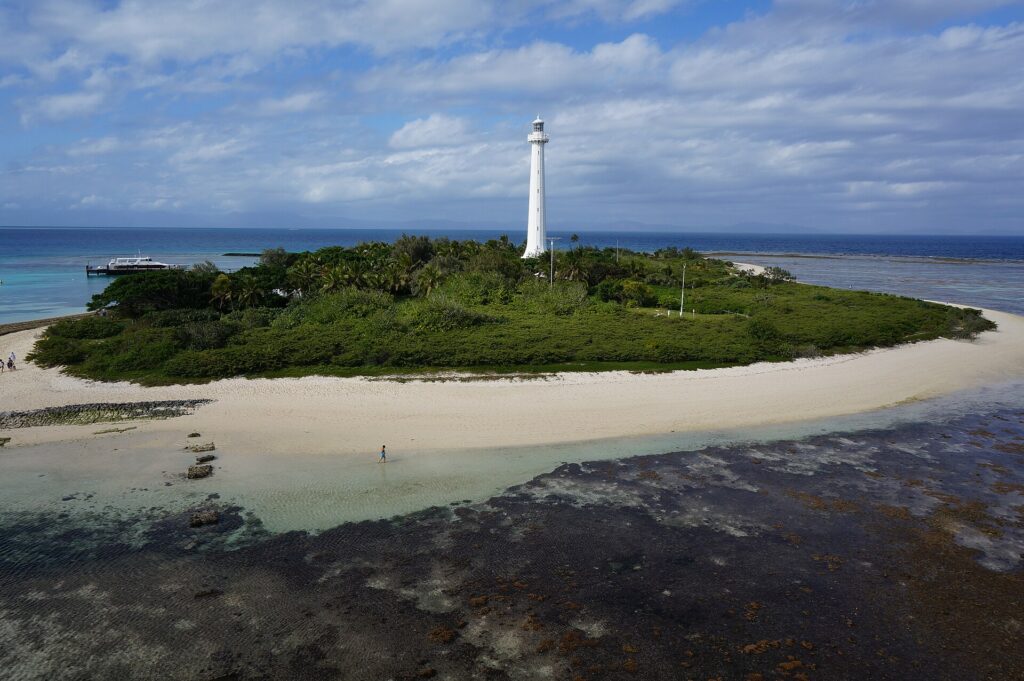 O grande farol branco da ilhota Amédée se ergue acima da vegetação, a ilhota cercada de areia branca, o platô de coral descoberto em primeiro plano e um barco atracado à esquerda