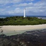 O grande farol branco da ilhota Amédée se ergue acima da vegetação, a ilhota cercada de areia branca, o platô de coral descoberto em primeiro plano e um barco atracado à esquerda