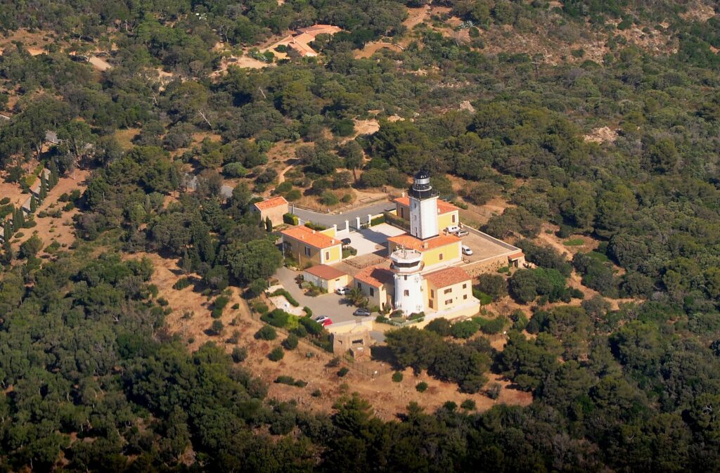 Vista aérea do farol do cabo Camarat, torre branca com lanterna preta cercada de prédios amarelos com telhados de telha laranja, no meio de uma densa floresta mediterrânea.