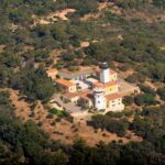 Vista aérea do farol do cabo Camarat, torre branca com lanterna preta cercada de prédios amarelos com telhados de telha laranja, no meio de uma densa floresta mediterrânea.