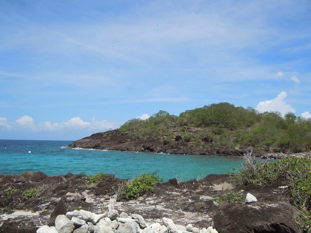 Uma enseada turquesa protegida por uma ponta de rocha escura coberta de mato, vista das rochas da ilhota, sob céu azul