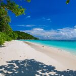 A praia de areia branca de Koh Tachai margeada de árvores sob um céu azul, com a água turquesa à direita e uma linha de boias ao largo