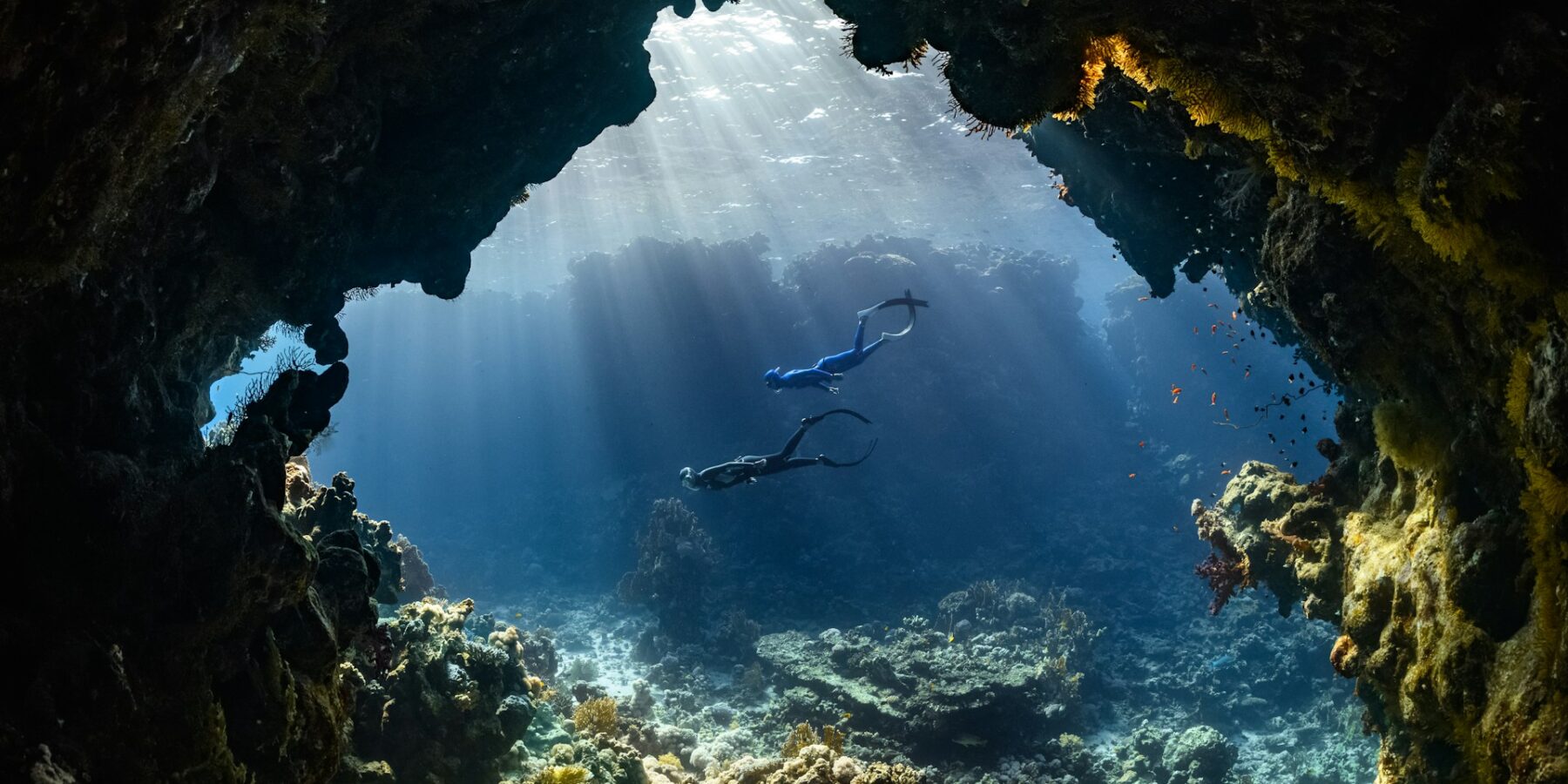 Dois mergulhadores atravessam uma caverna submarina iluminada pelos raios de sol, acima de um recife