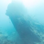 A popa partida do Thistlegorm, erguida e inclinada no azul, com um mergulhador à direita