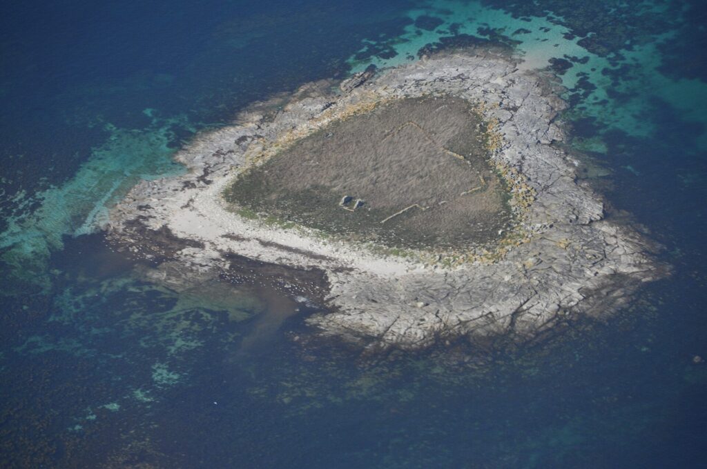 Vista aérea de uma pequena ilhota de lajes de granito cinza, coberta no centro de grama seca onde se adivinham muretas em ruínas, margeada por uma praia clara e cercada de baixios turquesa e de água azul profunda.