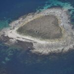 Vista aérea de uma pequena ilhota de lajes de granito cinza, coberta no centro de grama seca onde se adivinham muretas em ruínas, margeada por uma praia clara e cercada de baixios turquesa e de água azul profunda.