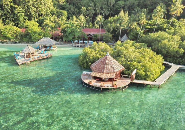 Vista aérea do Raja Ampat Dive Lodge, com píeres sobre palafitas e telhados vermelhos sob as palmeiras