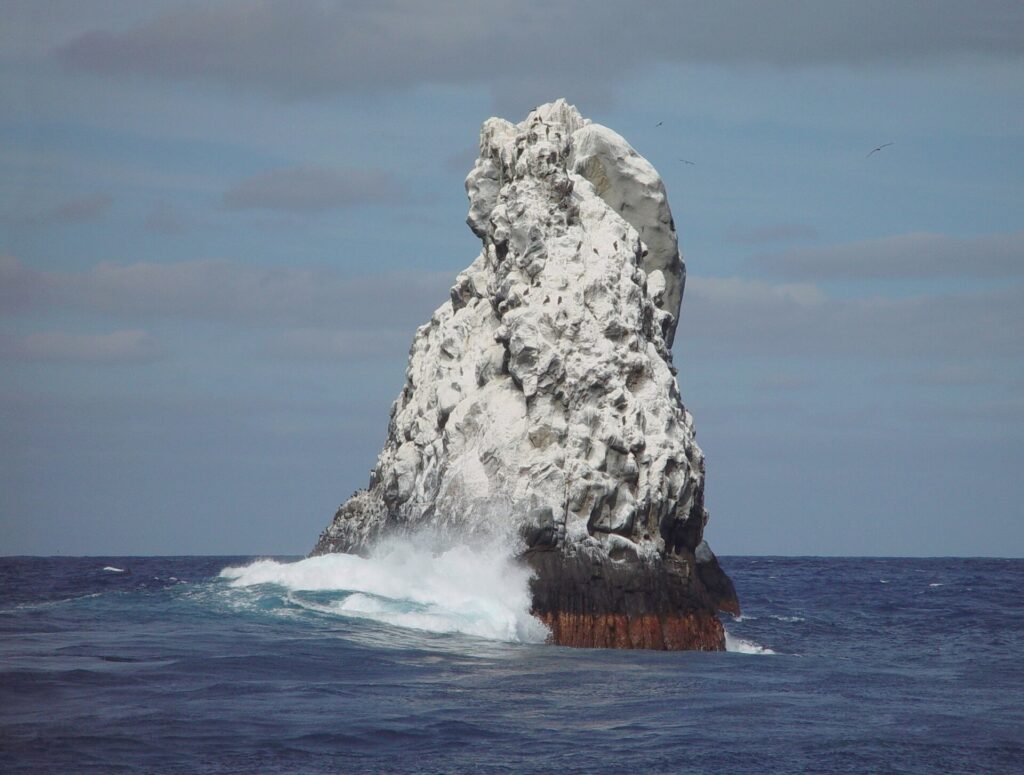 O pico noroeste de Roca Partida, branco de guano, visto do barco, com uma onda arrebentando na base e aves voando acima