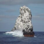 O pico noroeste de Roca Partida, branco de guano, visto do barco, com uma onda arrebentando na base e aves voando acima