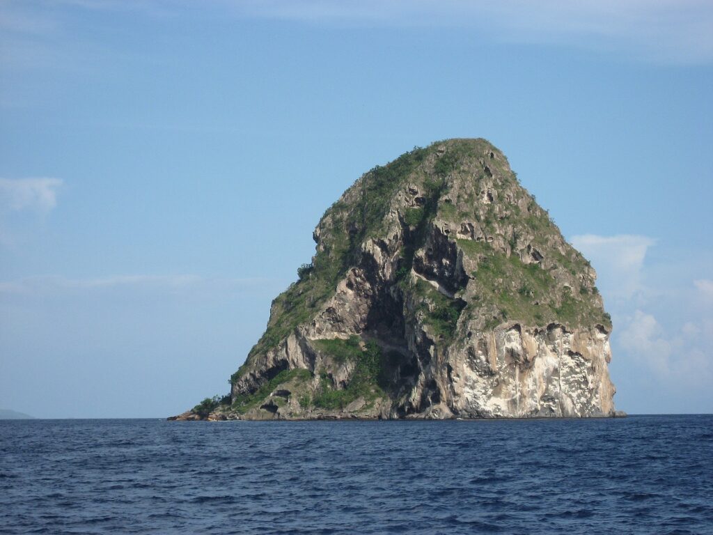 O Rochedo do Diamante visto de perto a partir do mar, uma massa de rocha cinza escavada de grutas, riscada de branco e coberta de vegetação no topo, acima de um mar azul-escuro