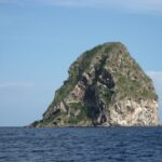 O Rochedo do Diamante visto de perto a partir do mar, uma massa de rocha cinza escavada de grutas, riscada de branco e coberta de vegetação no topo, acima de um mar azul-escuro