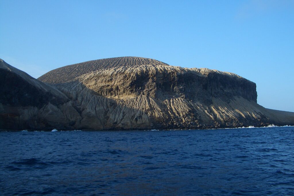 A costa de San Benedicto vista do mar: o cone de cinzas do vulcão Bárcena, riscado de ravinas, acima de falésias escuras e de um mar azul-escuro