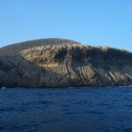 A costa de San Benedicto vista do mar: o cone de cinzas do vulcão Bárcena, riscado de ravinas, acima de falésias escuras e de um mar azul-escuro