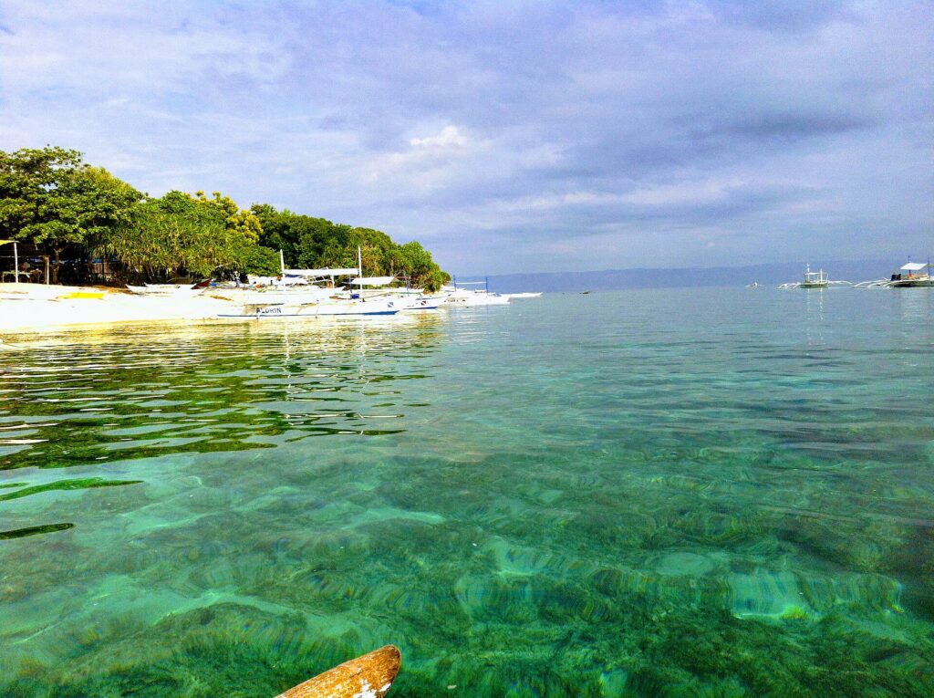 Água turquesa transparente sobre um fundo raso, diante de uma praia de areia branca margeada de árvores onde se enfileiram bangkas brancas, com outros barcos ao largo sob um céu nublado.