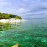 Água turquesa transparente sobre um fundo raso, diante de uma praia de areia branca margeada de árvores onde se enfileiram bangkas brancas, com outros barcos ao largo sob um céu nublado.