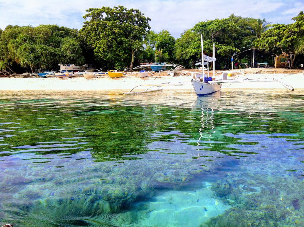 Rente à água, cabeços de coral sob uma água clara e turquesa, diante de uma praia de areia branca onde está ancorada uma bangka branca, com outros barcos coloridos puxados para debaixo das árvores.