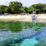 Rente à água, cabeços de coral sob uma água clara e turquesa, diante de uma praia de areia branca onde está ancorada uma bangka branca, com outros barcos coloridos puxados para debaixo das árvores.