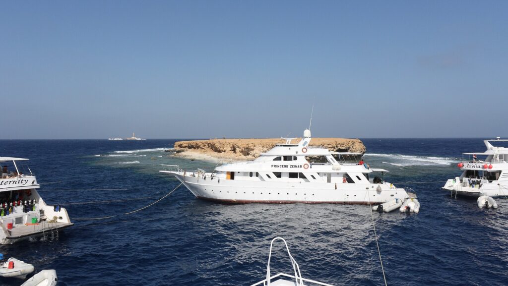 Três barcos de cruzeiro amarrados diante da ilhota plana de Small Brother, com o farol de Big Brother visível no horizonte