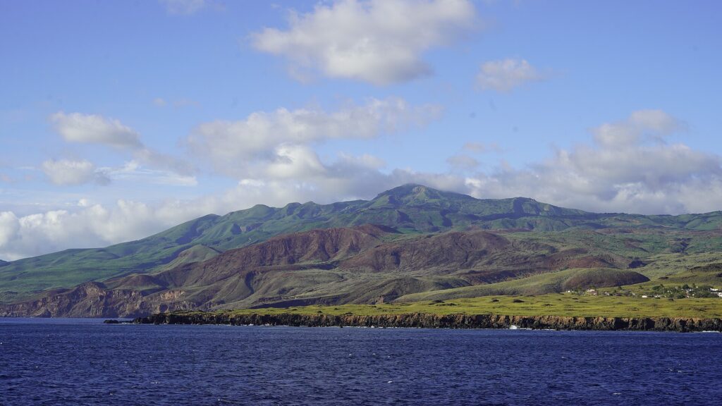 A costa da ilha Socorro vista do mar: falésias de lava negra, morros verdes e avermelhados e o topo do vulcão nas nuvens