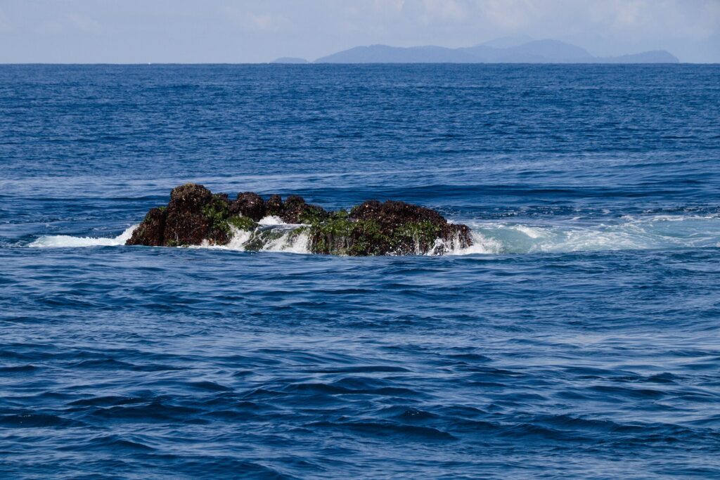 O topo de Richelieu Rock aparece na maré baixa, uma rocha escura coberta de algas verdes e batida pelas ondas, com as ilhas Surin no horizonte