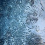 Um cardume cerrado de sardinhas prateadas roça um fundo de areia muito clara salpicado de rochas escuras, com a massa de peixes mais densa à esquerda, no azul.