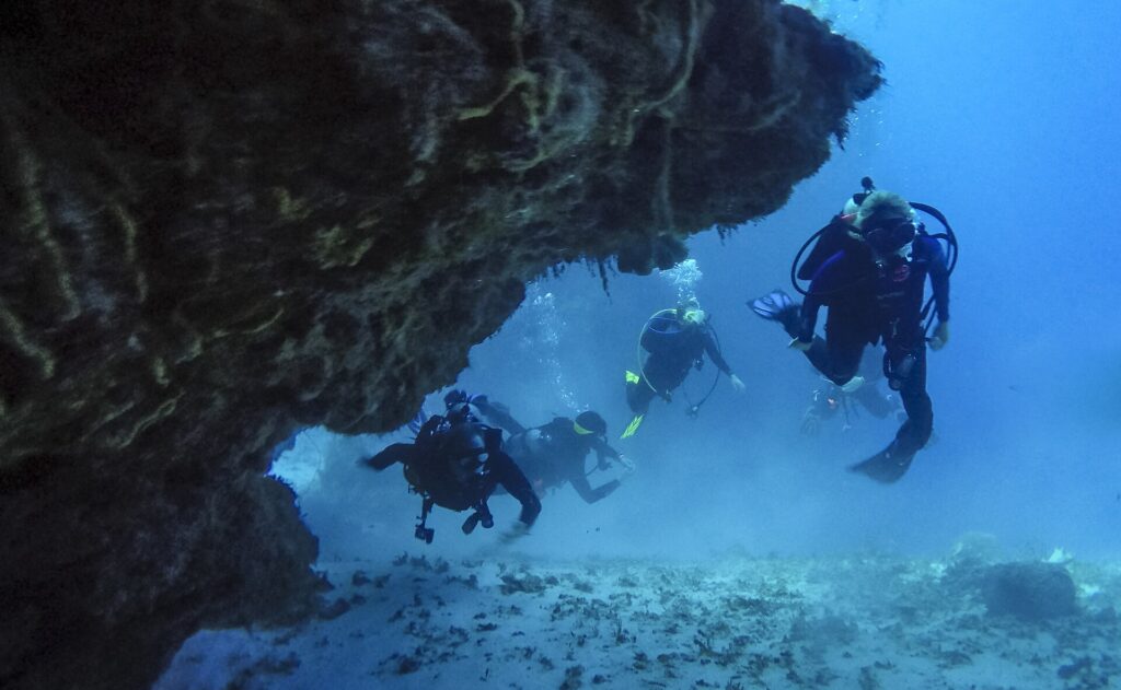 Quatro mergulhadores saem de um túnel de coral sobre um fundo de areia, em Cozumel, com o teto de rocha acima deles