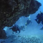 Quatro mergulhadores saem de um túnel de coral sobre um fundo de areia, em Cozumel, com o teto de rocha acima deles