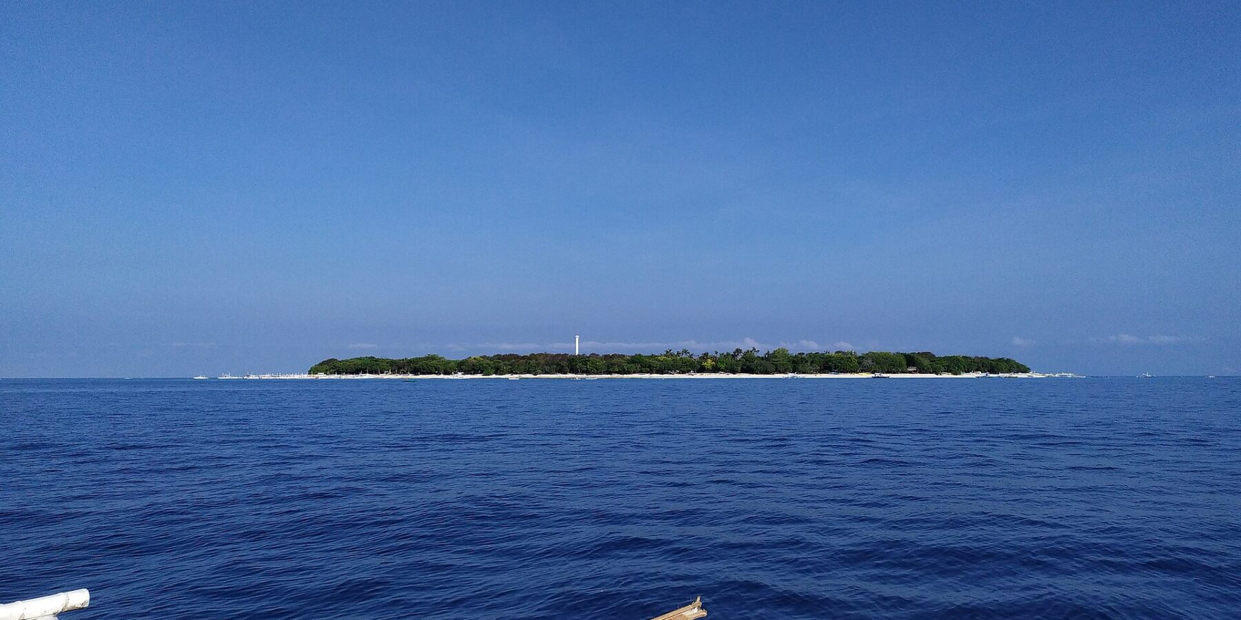 A ilha plana e arborizada de Balicasag no horizonte, com o farol branco e a faixa de areia clara, vista de uma bangka cujos flutuadores brancos ocupam o primeiro plano, sob um céu azul sem nuvens.