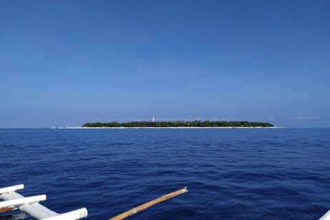 A ilha plana e arborizada de Balicasag no horizonte, com o farol branco e a faixa de areia clara, vista de uma bangka cujos flutuadores brancos ocupam o primeiro plano, sob um céu azul sem nuvens.