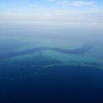 Vista aérea de um canal azul-escuro em forma de S que serpenteia entre os platôs turquesa da barreira de coral, com alguns barquinhos brancos no canal