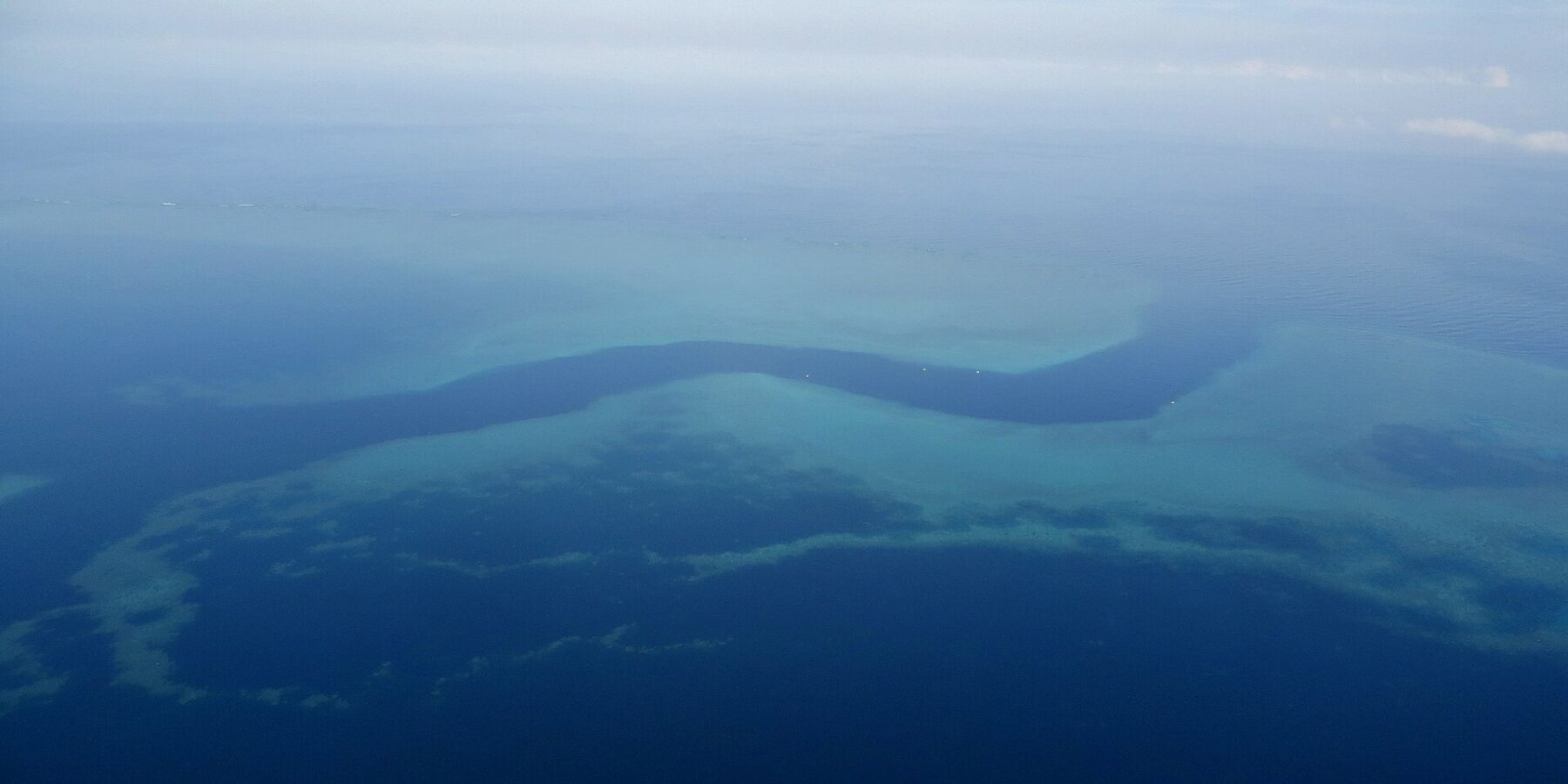 Vista aérea de um canal azul-escuro em forma de S que serpenteia entre os platôs turquesa da barreira de coral, com alguns barquinhos brancos no canal
