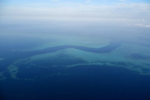 Vista aérea de um canal azul-escuro em forma de S que serpenteia entre os platôs turquesa da barreira de coral, com alguns barquinhos brancos no canal