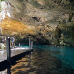 O olho leste do cenote Dos Ojos: uma abóbada de calcário eriçada de estalactites sobre uma água turquesa, com um deque de madeira à esquerda e banhistas ao fundo
