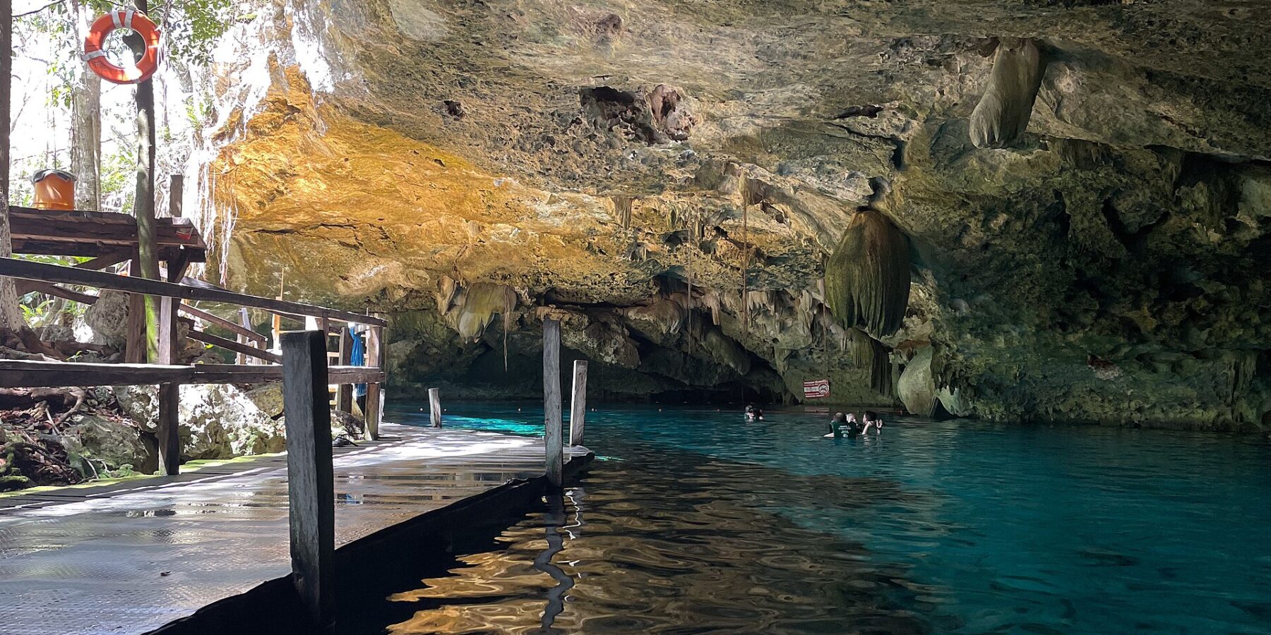 O olho leste do cenote Dos Ojos: uma abóbada de calcário eriçada de estalactites sobre uma água turquesa, com um deque de madeira à esquerda e banhistas ao fundo