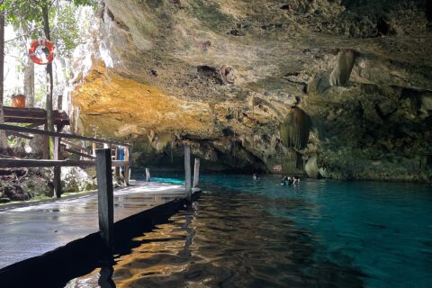 O olho leste do cenote Dos Ojos: uma abóbada de calcário eriçada de estalactites sobre uma água turquesa, com um deque de madeira à esquerda e banhistas ao fundo