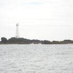 Um farol branco e esguio de porta vermelha se ergue na ilhota baixa e rochosa de Formigão, com rochas escuras orladas de algas marrons, acima de um mar cinza e agitado sob um céu encoberto