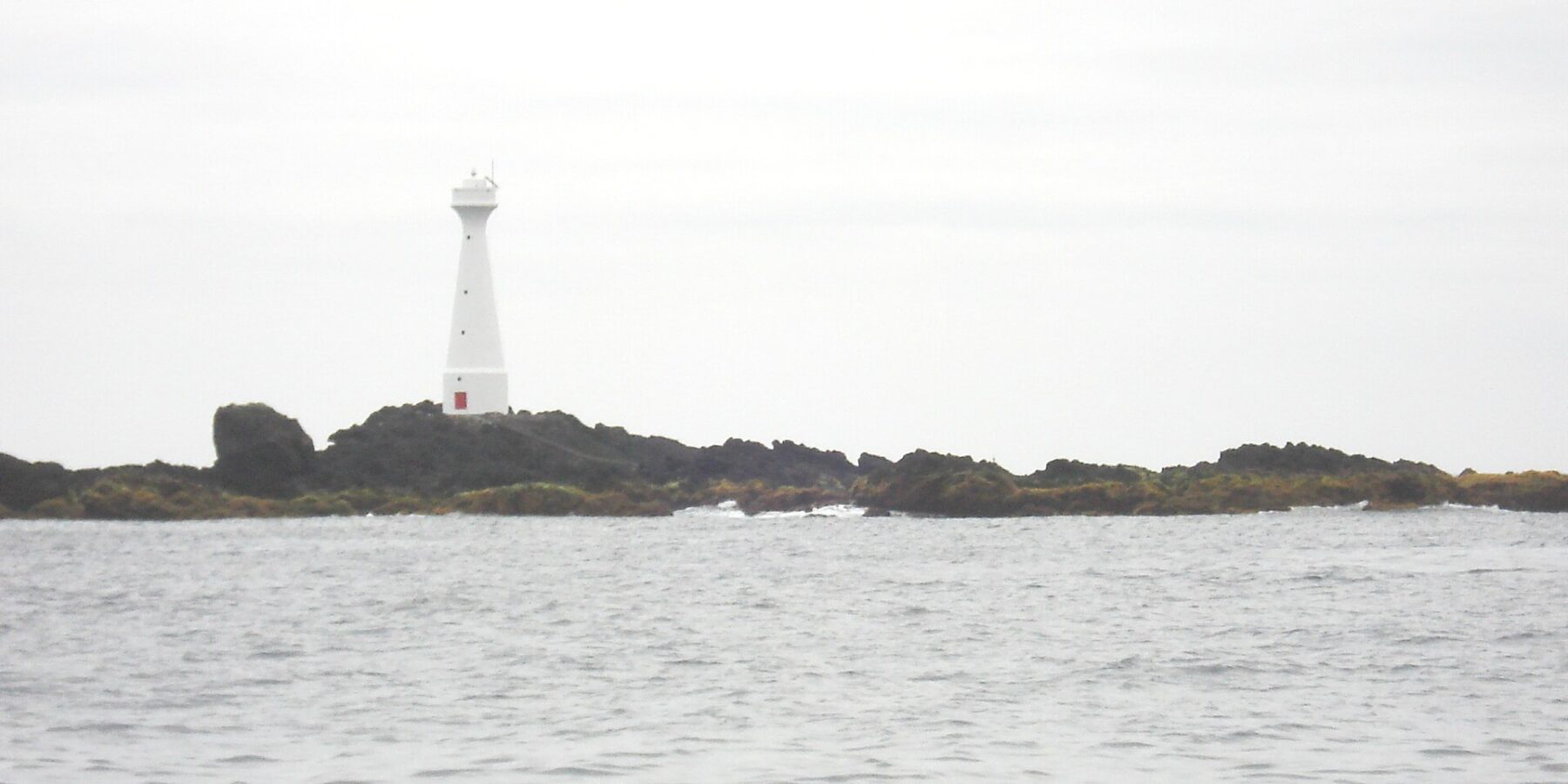 Um farol branco e esguio de porta vermelha se ergue na ilhota baixa e rochosa de Formigão, com rochas escuras orladas de algas marrons, acima de um mar cinza e agitado sob um céu encoberto