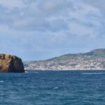 Panorama dos dois ilhéus de tufo ocre da Madalena num mar azul-escuro, o Ilhéu Deitado comprido e plano à esquerda e o Ilhéu em Pé erguido à direita, com a cidade branca da Horta e as colinas verdes do Faial ao fundo