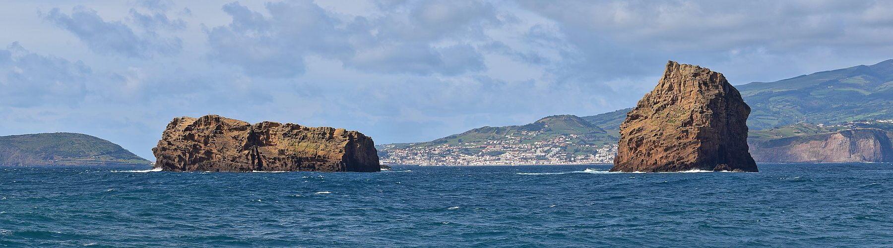 Panorama dos dois ilhéus de tufo ocre da Madalena num mar azul-escuro, o Ilhéu Deitado comprido e plano à esquerda e o Ilhéu em Pé erguido à direita, com a cidade branca da Horta e as colinas verdes do Faial ao fundo