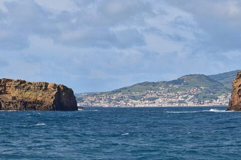 Panorama dos dois ilhéus de tufo ocre da Madalena num mar azul-escuro, o Ilhéu Deitado comprido e plano à esquerda e o Ilhéu em Pé erguido à direita, com a cidade branca da Horta e as colinas verdes do Faial ao fundo