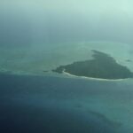 A ilha de Misali vista de avião, um triângulo de floresta margeado de praia branca, sobre um platô recifal turquesa no meio de um mar azul-escuro, na bruma