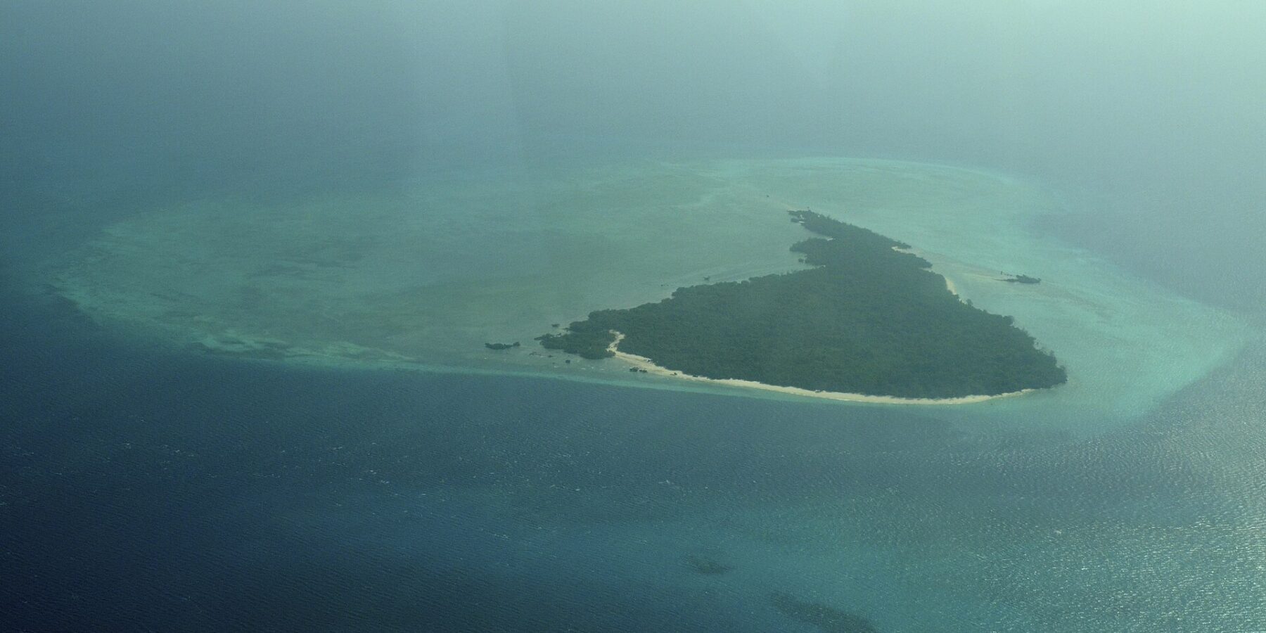 A ilha de Misali vista de avião, um triângulo de floresta margeado de praia branca, sobre um platô recifal turquesa no meio de um mar azul-escuro, na bruma