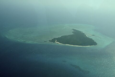 A ilha de Misali vista de avião, um triângulo de floresta margeado de praia branca, sobre um platô recifal turquesa no meio de um mar azul-escuro, na bruma