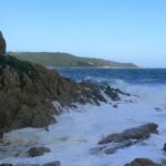 Ondas se quebram em espuma branca nas rochas de granito de L'Escalet, em primeiro plano, diante de um mar azul-escuro agitado, com a costa arborizada do cabo Camarat e seu farol branco ao longe.
