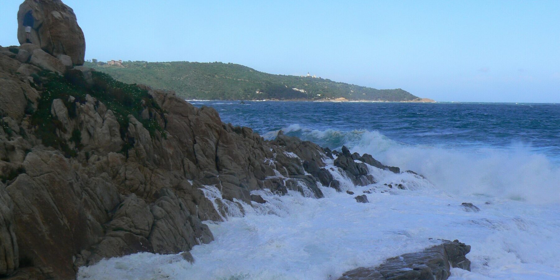 Ondas se quebram em espuma branca nas rochas de granito de L'Escalet, em primeiro plano, diante de um mar azul-escuro agitado, com a costa arborizada do cabo Camarat e seu farol branco ao longe.