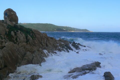 Ondas se quebram em espuma branca nas rochas de granito de L'Escalet, em primeiro plano, diante de um mar azul-escuro agitado, com a costa arborizada do cabo Camarat e seu farol branco ao longe.