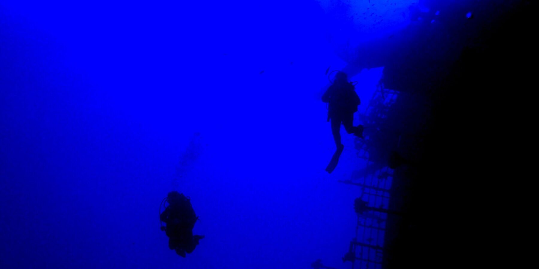 No azul profundo, dois mergulhadores em silhueta passam ao longo da amurada da popa do Salem Express, com o convés inclinado à direita